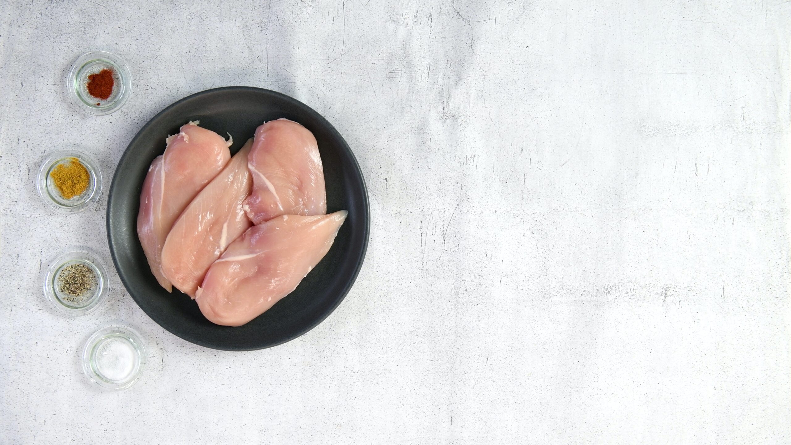 A plate with four chicken breasts on it, with 4 small bowls with salt, paprika and other seasonings in them.